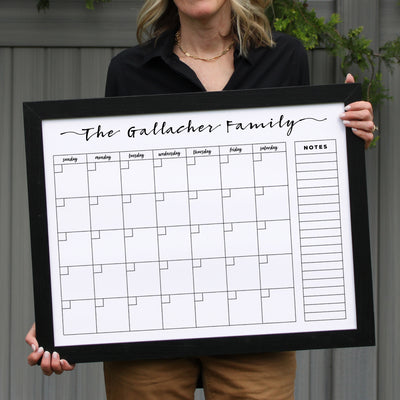 A person confidently stands against a gray wall holding the 24 X 18 Framed Whiteboard Calendar (#18203) by CalendarGirlCompany, labeled The Gallacher Family. The calendar includes a weekly grid and notes section, ideal for chalk ink markers. The individual is dressed in a black top and beige pants.