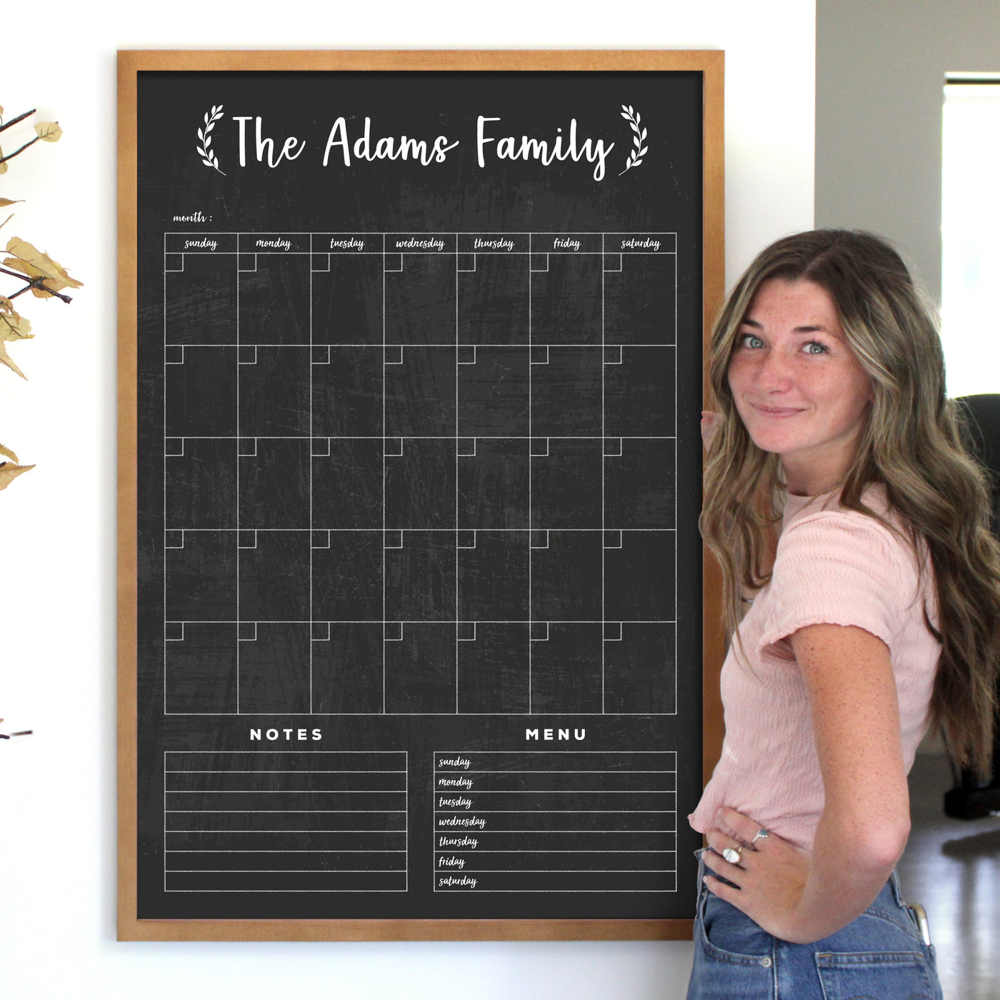 A woman with long hair stands smiling beside a CalendarGirlCompany 24 x 36 Large Vertical Chalkboard Calendar (#24131) labeled The Adams Family. The sleek black design with white text includes sections for days, notes, and menus. She wears a light pink shirt and uses chalk ink markers to organize everything.