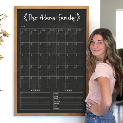 A woman with long hair stands smiling beside a CalendarGirlCompany 24 x 36 Large Vertical Chalkboard Calendar (#24131) labeled The Adams Family. The sleek black design with white text includes sections for days, notes, and menus. She wears a light pink shirt and uses chalk ink markers to organize everything.