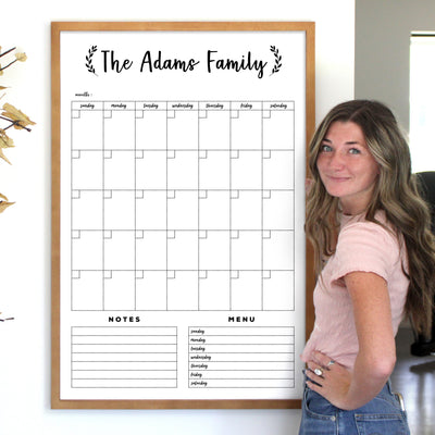A woman stands beside the 24 x 36 LARGE VERTICAL WHITEBOARD FRAMED CALENDAR #24131 by CalendarGirlCompany, with The Adams Family elegantly at the top. Its premium acrylic surface has daily sections for notes and a menu. Smiling warmly in her light pink shirt, shes ready to enjoy free customizations.