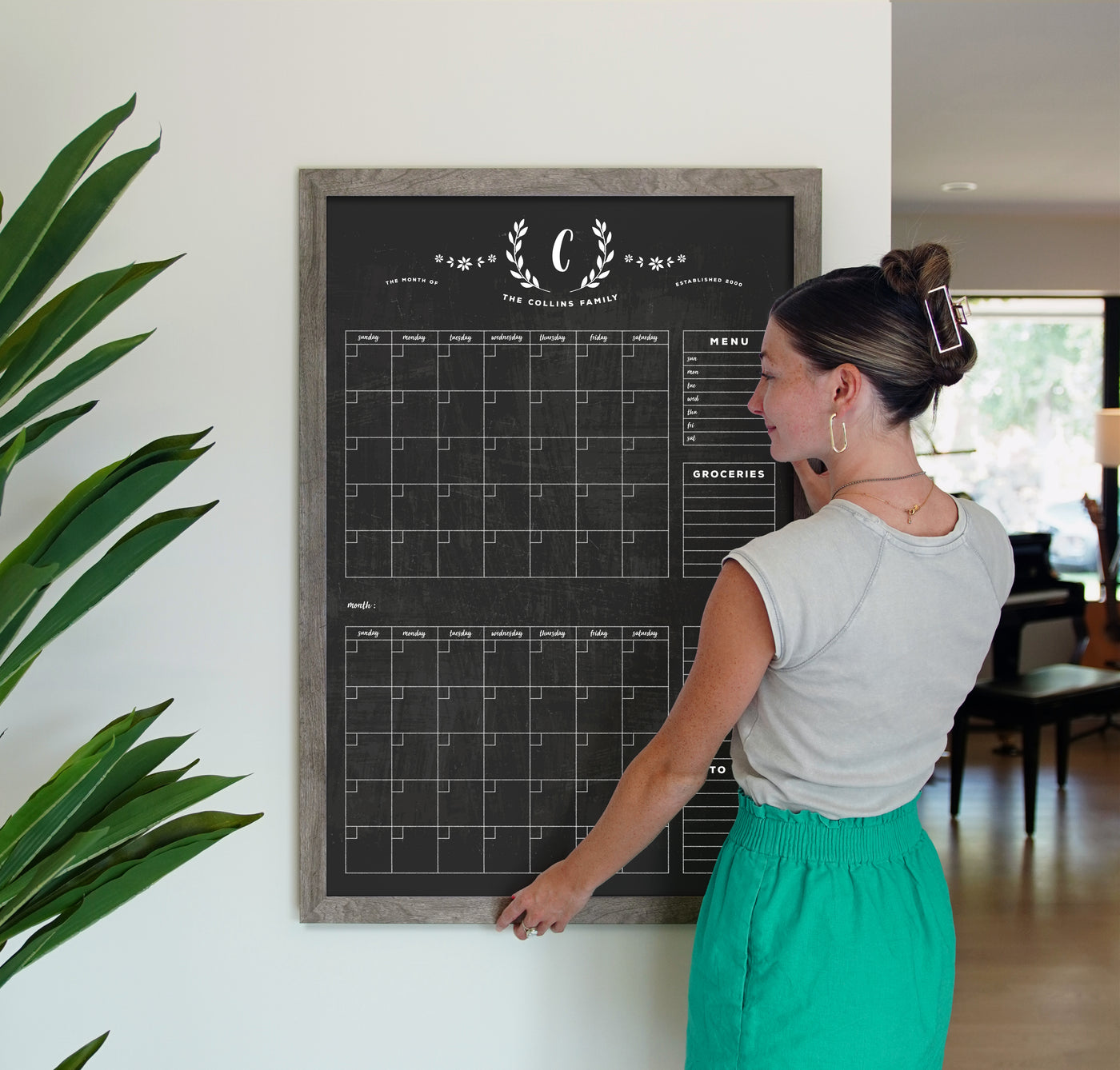 A woman in a gray top and green skirt writes with chalk ink markers on the 24 x 36 LARGE VERTICAL CHALKBOARD - 24132 by CalendarGirlCompany in a well-lit room. A tall green plant is to her left, while sunlight filters through the window.