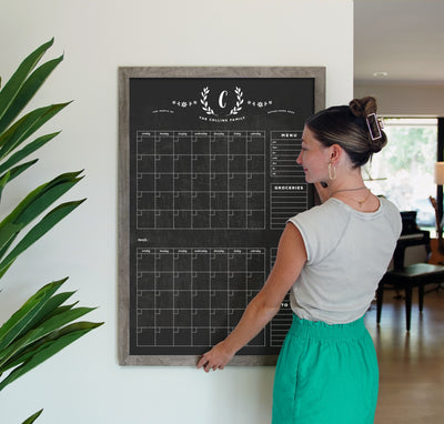 A woman in a gray top and green skirt writes with chalk ink markers on the 24 x 36 LARGE VERTICAL CHALKBOARD - 24132 by CalendarGirlCompany in a well-lit room. A tall green plant is to her left, while sunlight filters through the window.