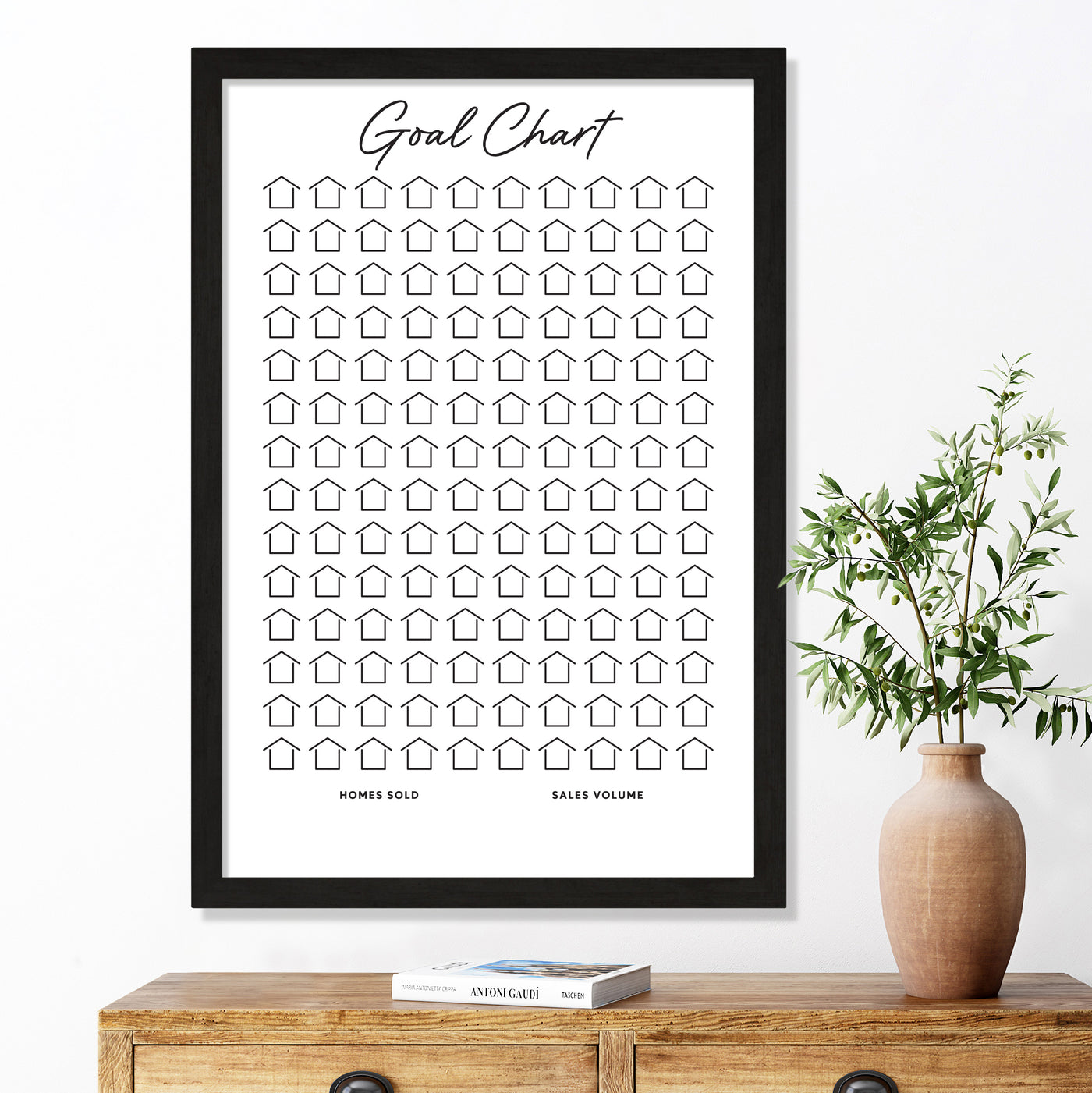 The Girl Friday Shops Framed Goal Chart features 100 house icons labeled Homes Sold and Sales Volume, set above a wooden table with a potted plant and two books, enhanced by a premium acrylic finish. This visual Sales Tracker for Real Estate adds elegance to your space.
