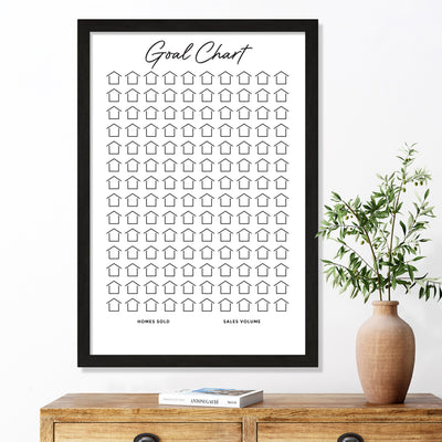 The Girl Friday Shops Framed Goal Chart features 100 house icons labeled Homes Sold and Sales Volume, set above a wooden table with a potted plant and two books, enhanced by a premium acrylic finish. This visual Sales Tracker for Real Estate adds elegance to your space.