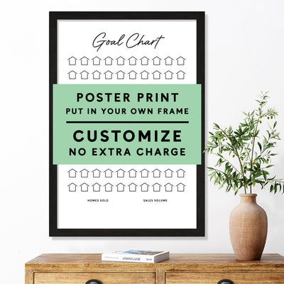 A framed Goal Chart Poster Print by Girl Friday Shop sits on a wooden table with a potted plant. It features rows of house icons and a customizable text section.