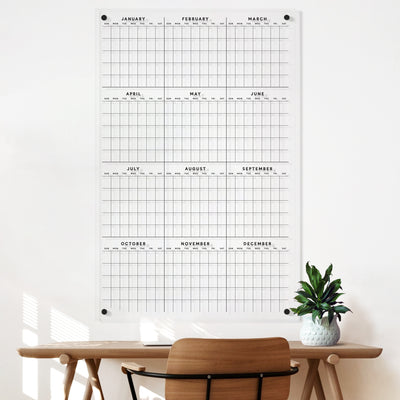 The Girl Friday Clear Acrylic Yearly Wall Calendar, with polished edges and a grid format for months and days, shines in the sunlight of this bright room. A wooden chair and desk with a small potted plant add to the serene scene.