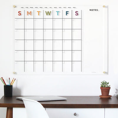 A minimalistic office setup includes a wooden desk with a closed laptop, colored pencils in a cup, and a small potted plant. On the wall, the Girl Friday Acrylic Calendar EARTH TONE RAINBOW displays colorful letters representing days of the week.