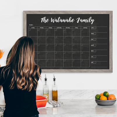 In a kitchen with marble counters hosting olive oil, vinegar, a red dish, and fruit, a woman writes on a CalendarGirlCompanys #24126 36 x 24 Framed Chalkboard Calendar using vibrant chalk ink markers. The calendar is labeled The Watanabe Family and mounted on a white wall in the background.