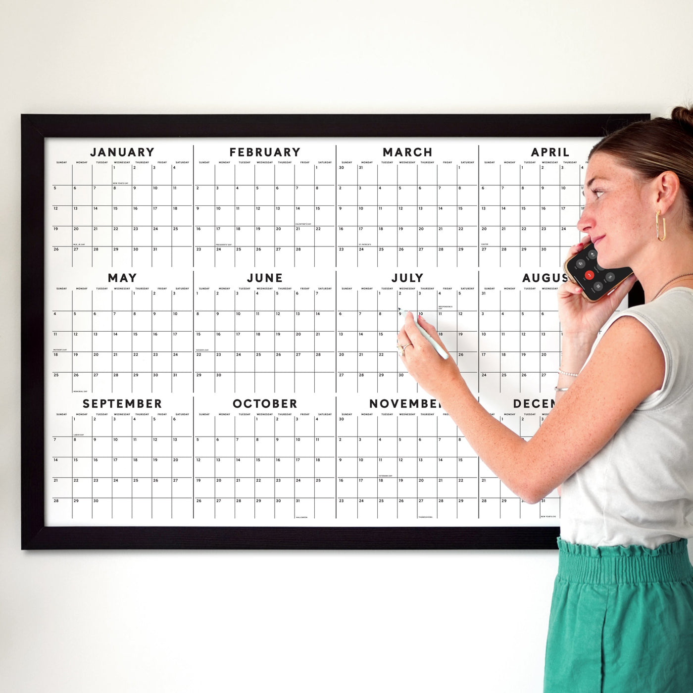 A woman in a white tank top and green pants talks on her phone, pointing at the month of July on a framed Girl Friday 2025 Yearly Calendar with Dates and Holidays.