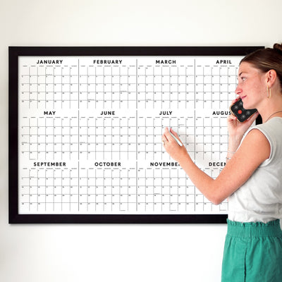 A woman in a white tank top and green pants talks on her phone, pointing at the month of July on a framed Girl Friday 2025 Yearly Calendar with Dates and Holidays.