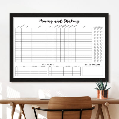 The Girl Friday Real Estate Moving & Shaking Whiteboard with customizable frame options hangs above a wooden desk and chair, featuring sections for listings, sales goals, hot leads, and sales volume. A small potted plant sits nearby.
