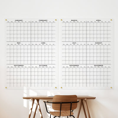 A minimalist workspace boasts a wooden chair and desk. Two Girl Friday yearly dry-erase acrylic calendars are mounted on the wall using discreet hardware, featuring clear polished edges. Sunlight casts shadows of the chair and desk onto the pristine white wall.