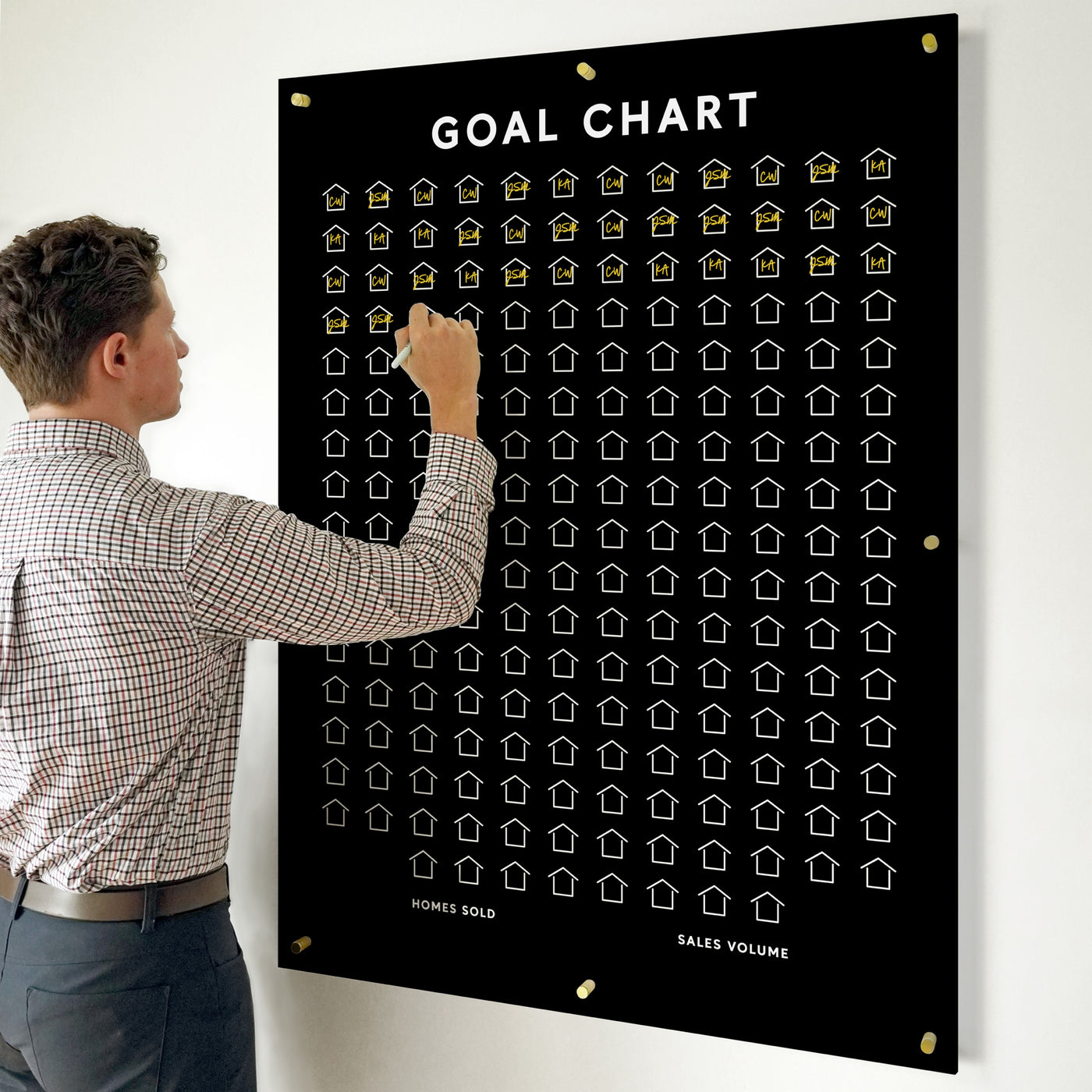 A man updates a Girl Friday Goal Chart | Sales Tracker for any business on black acrylic, featuring rows of house icons. Some icons shine in gold, indicating progress, while the title Goal Chart is prominently displayed at the top.