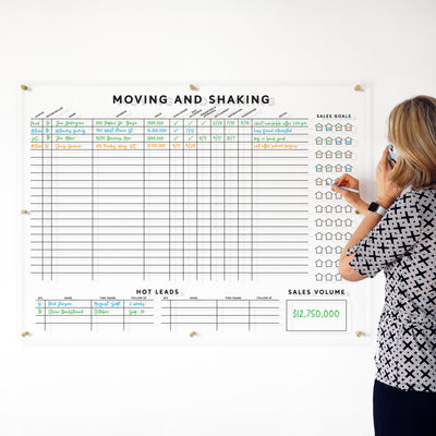 A person stands before a wall-mounted Moving & Shaking Clear Acrylic Real Estate Board by Girl Friday, displaying a grid with sales data, goals, and hot leads. They write with a marker while holding a phone to their ear.