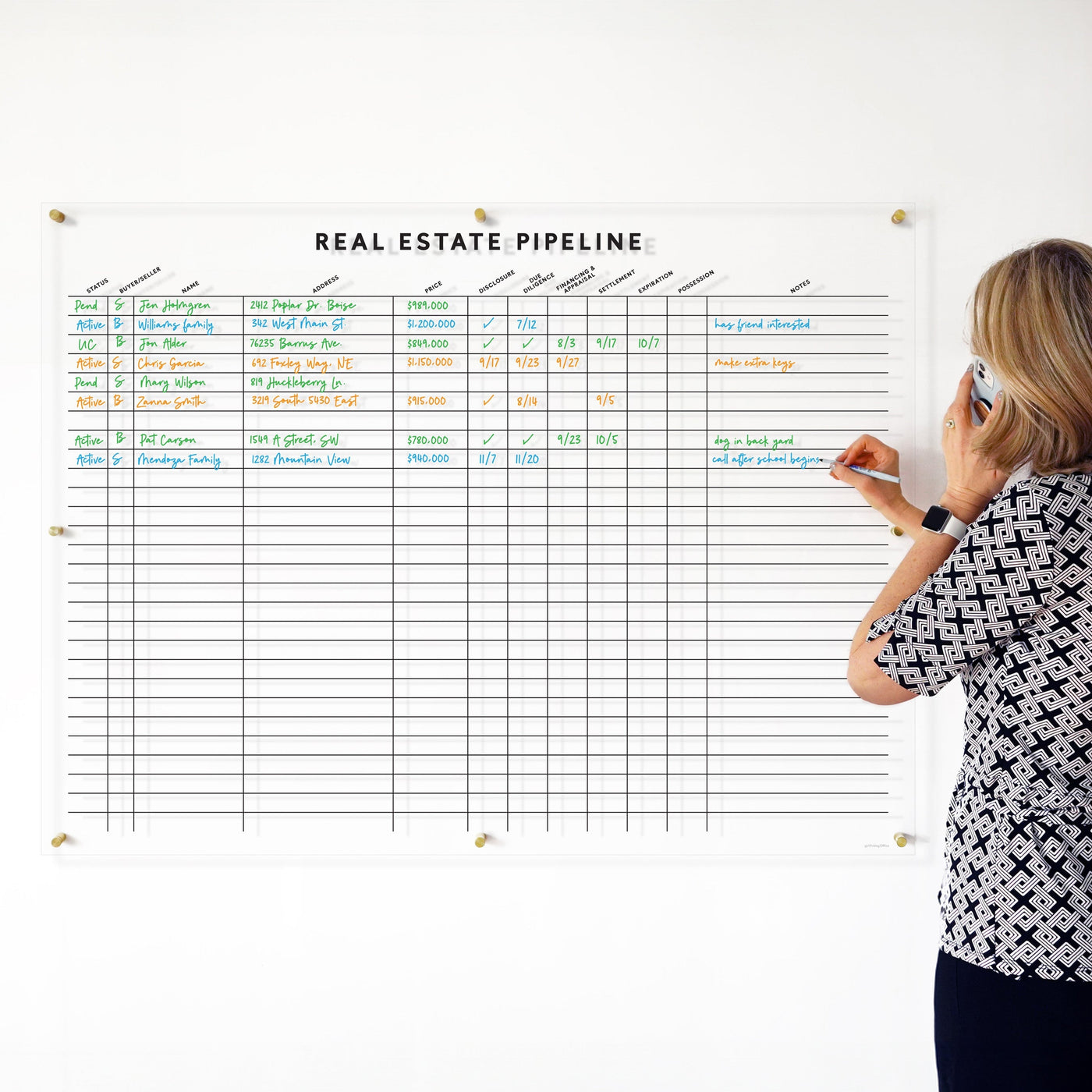 A woman chats on the phone, writing on her customized Girl Friday Real Estate Pipeline Acrylic Board, showcasing property details, financials, and status updates in vibrant wet-erase marker colors against a sleek white backdrop.