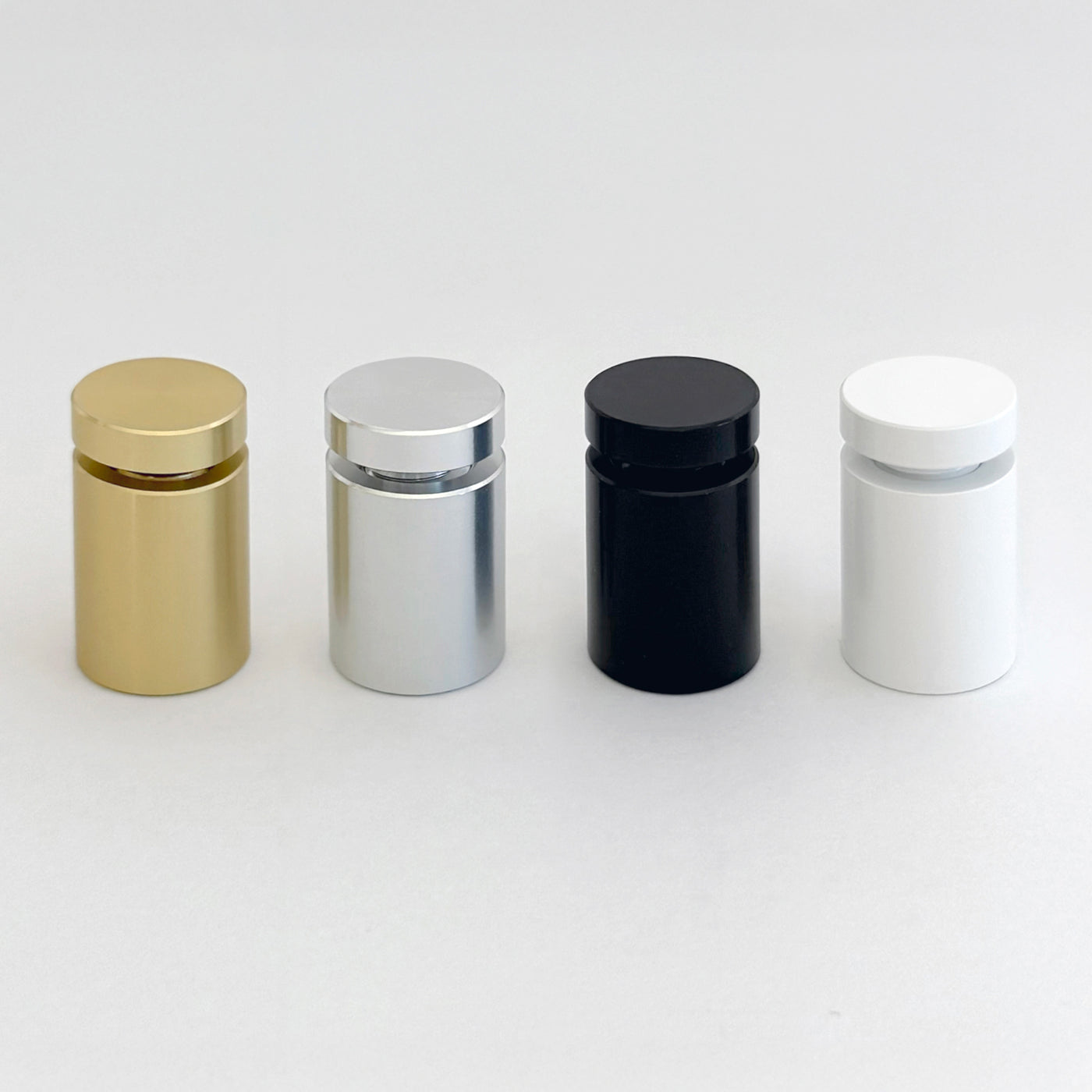 Four Modern Corkboard bottles by Girl Friday Shop sit on a white surface, offering a floating appearance. Each cylindrical bottle has a rounded cap and comes in gold, silver, black, or white with a metallic sheen and minimalist design.
