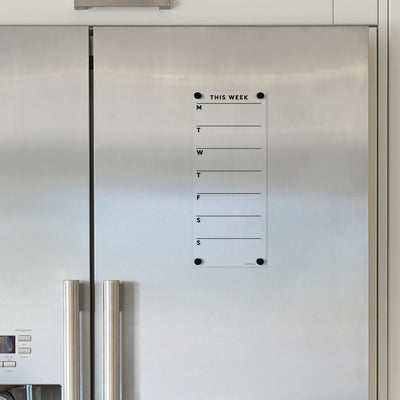 A Girl Friday stainless steel refrigerator featuring the Clear Acrylic Fridge Planner with a customizable title and black text. It boasts double doors, long handles, and includes a weekly planner (Monday to Sunday) for use with wet-erase markers, offering style and convenience.