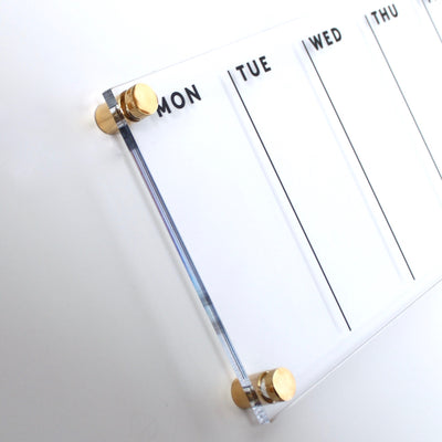 The Acrylic Calendar - weekly by Girl Friday Shop is a high-quality organizer featuring a clear planner with gold standoffs for Monday to Thursday. Perfectly mounted on a white wall, its designed for use with a wet-erase marker.