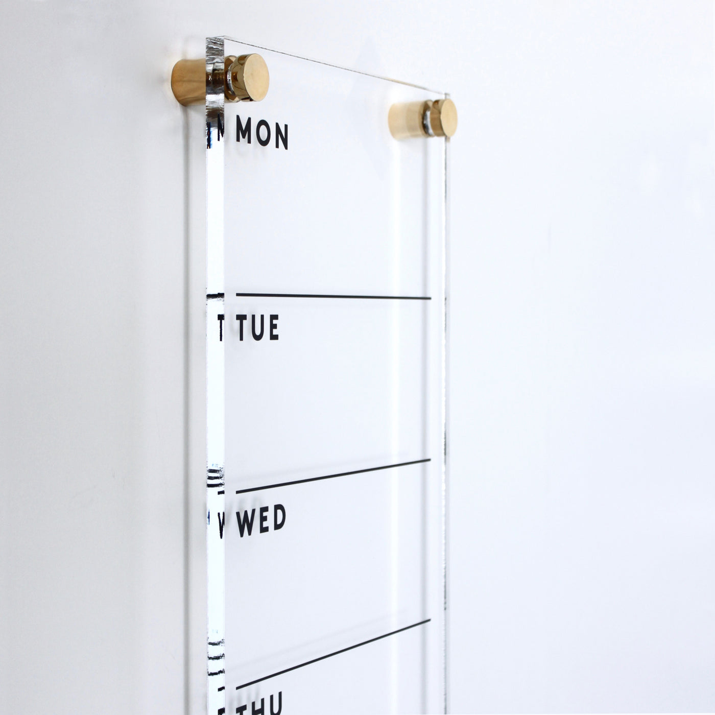 The Girl Friday Shops Acrylic Calendar - weekly is a clear organizer mounted on a white wall, featuring Monday to Thursday with note lines. Made from high-quality materials and held by two wooden pegs, it pairs perfectly with a wet-erase marker.