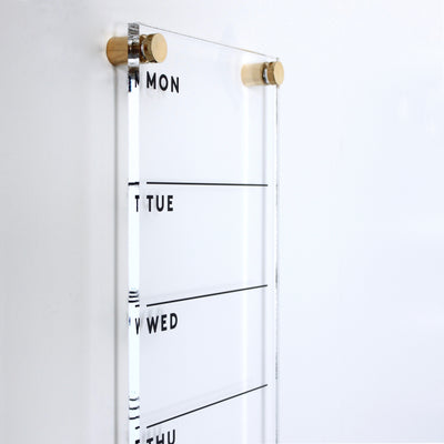 The Girl Friday Shops Acrylic Calendar - weekly is a clear organizer mounted on a white wall, featuring Monday to Thursday with note lines. Made from high-quality materials and held by two wooden pegs, it pairs perfectly with a wet-erase marker.