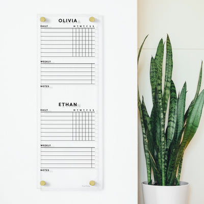 The Girl Friday vertical Clear Acrylic Customizable Chore Chart, designed for 1-5 children with sections like Olivia and Ethan, features white spaces for notes and gold pegs. It contrasts beautifully with a tall green plant and is perfect for dry-erase markers.