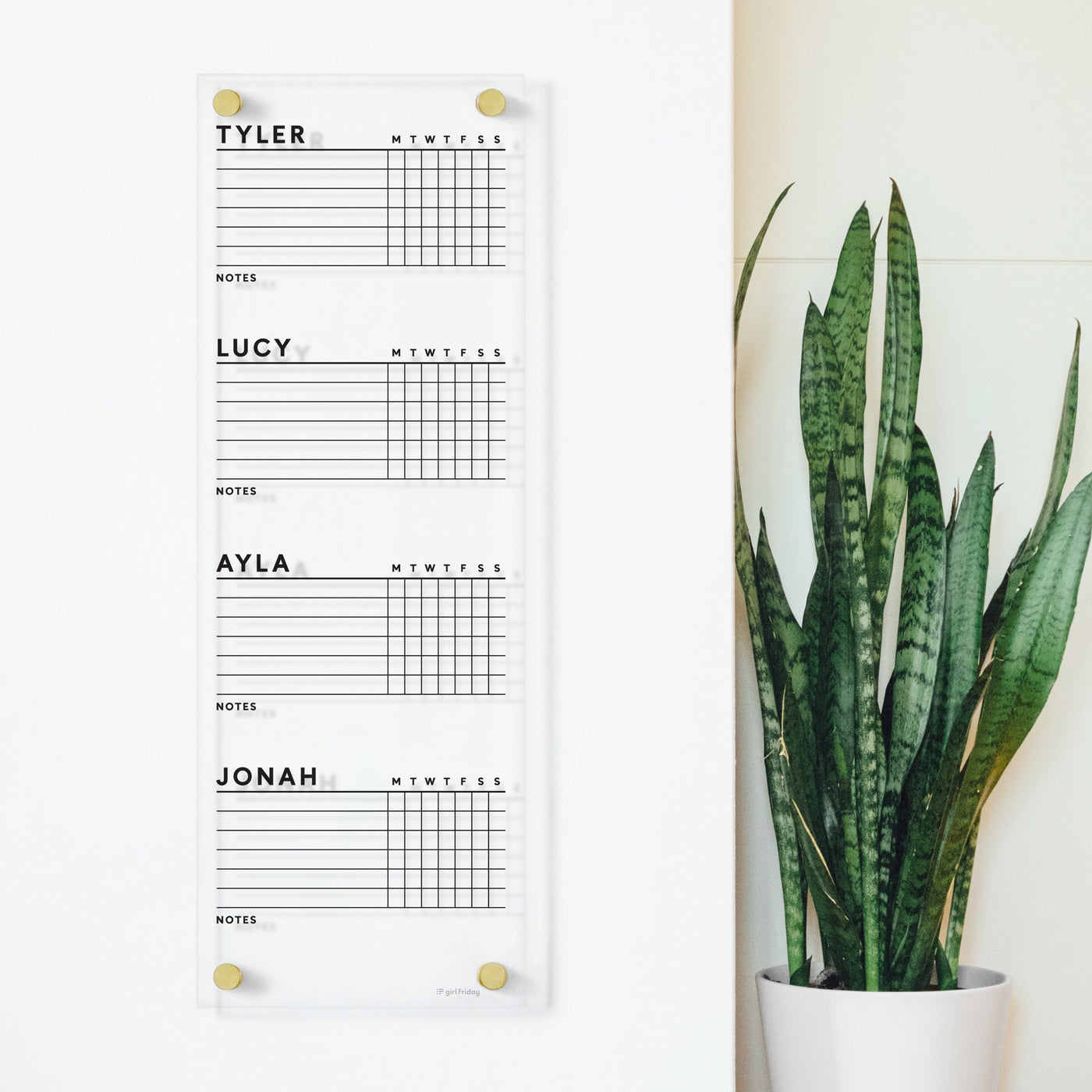 The Girl Friday Clear Acrylic Customizable Chore Chart, ideal for 1-5 children, is wall-mounted beside a tall green plant. It features sections for Tyler, Lucy, Ayla, and Jonah with days of the week and a notes area. Perfectly compatible with dry-erase markers for easy updates.