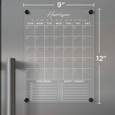 The Girl Friday Clear Acrylic Monthly Fridge Calendar includes customizable bottom sections with white text, featuring columns for each day and spaces for notes labeled This Week and Dont Forget. It measures 9x12 inches and is designed for wet-erase markers.