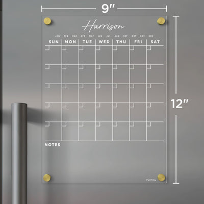 The Harrison by Girl Friday is a 9x12-inch clear acrylic wall calendar with a Sunday-start grid layout, note space at the bottom, and gold corner mounts. Its compatible with dry-erase markers and perfect for home office or fridge use.