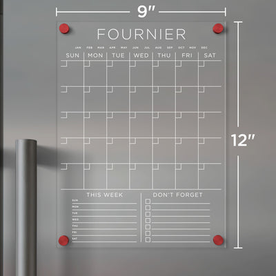 The Girl Friday Clear Acrylic Monthly Fridge Calendar features a grid layout and customizable family name. With sections titled THIS WEEK and DONT FORGET, its ideal for wet-erase markers. A pin system secures it to your fridge, dimensions 9 x 12. White text design on an acrylic board.