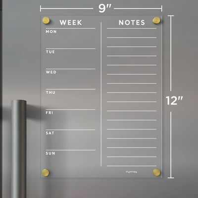 The Girl Friday Clear Acrylic Customizable Fridge Calendar, featuring gold corners and measuring 9x12 inches, is shown on a gray metal surface. It offers daily sections on the left and a notes area on the right, ideal for wet-erase markers.