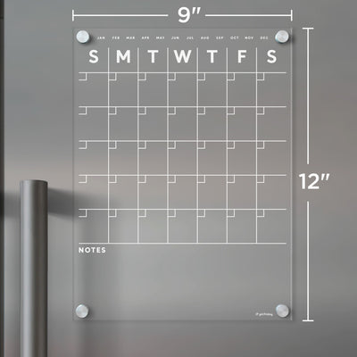 The Clear Acrylic Monthly Fridge Calendar by Girl Friday Shop features a transparent grid, bottom note space, and white text. It includes days and monthly markers from January to December. Measuring 9 x 12, its magnetic surface is ideal for wet-erase markers.