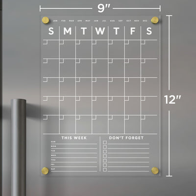 The Girl Friday Shop Clear Acrylic Monthly Fridge Calendar, measuring 9x12 inches, attaches to metallic surfaces with gold magnets. It features S-M-T-W-T-F-S daily boxes, This Week and Don’t Forget sections, customizable areas at the bottom, and is compatible with dry-erase markers.