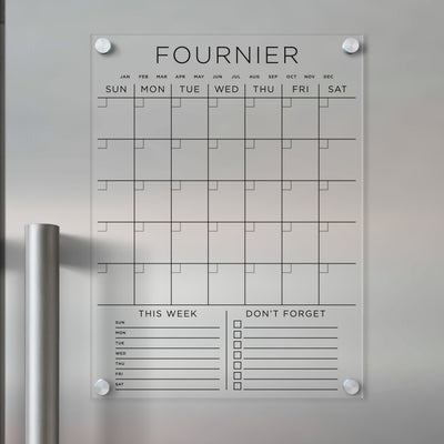A Girl Friday Acrylic Fridge Calendar with a custom family name is displayed on a silver fridge. It features sections titled This Week and Dont Forget, complete with checkboxes. Ideal for wet-erase markers, it keeps schedules visible and organized.