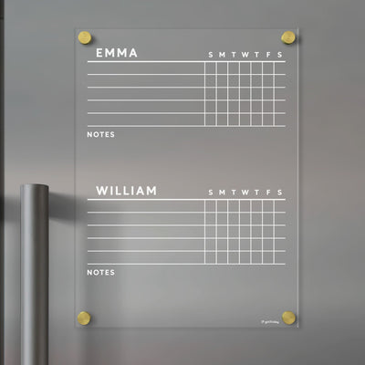 A sleek Girl Friday Acrylic Fridge Chore Chart for 2 children, featuring Emma and William sections with weekly planners and note space, is mounted on a gray wall. Elegantly enhanced by round gold fixtures, its ideal for organizing with wet-erase markers.