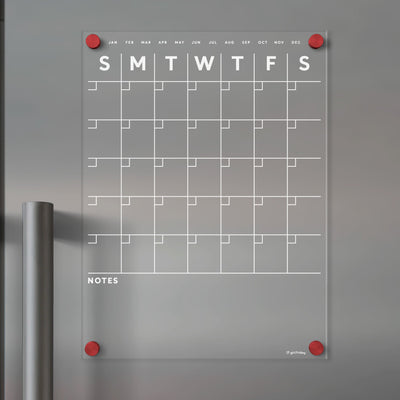The Acrylic Fridge Calendar by Girl Friday features white text and a transparent acrylic design. Its wall-mounted with red pegs, ideal for dry-erase markers, displaying week days and months, with a NOTES section under the grid against a gray background.