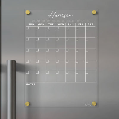 The Clear Acrylic Monthly Fridge Calendar by Girl Friday features a minimalist design with white outlines for wet-erase markers. Harrison is custom at the top, NOTES at the bottom, and gold fasteners in each corner. It showcases days of the week with large date squares.