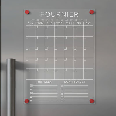 The Clear Acrylic Monthly Fridge Calendar by Girl Friday features daily columns, monthly rows, and sections labeled This Week and Dont Forget with checkboxes. Customizable with your family name, its perfect for wet-erase markers and is attached using red pins.