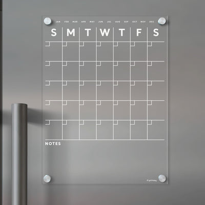 The Girl Friday Shops Clear Acrylic Monthly Fridge Calendar features a grid layout for days and months, with a bottom notes section. Its wall-mounted using visible screws and offers a modern, minimalist design. Use wet-erase markers for easy updates on this sleek surface.