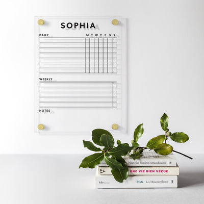 A Clear Acrylic Customizable Chore Chart by Girl Friday is mounted on the wall under the name Sophia, with sections for daily tasks, weekly tasks, and notes. Designed for wet-erase markers. Below, three stacked books hold a green plant branch.