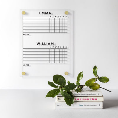 The Clear Acrylic Customizable Chore Chart by Girl Friday, perfect for 1-5 children, features EMMA and WILLIAM labels. This vertical wall-mounted planner has columns for each day and comes with a stack of books like Nouvelles histoires extraordinaires under a leafy branch. Use dry-erase markers to stay dynamic.