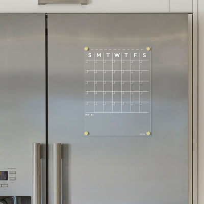 The Girl Friday Shop Clear Acrylic Monthly Magnetic Fridge Calendar, with white text, attaches to a stainless steel refrigerator. It features a minimalist design, empty daily squares from Sunday to Saturday, and a bottom notes section, ideal for use with a wet-erase marker on its magnetic surface.