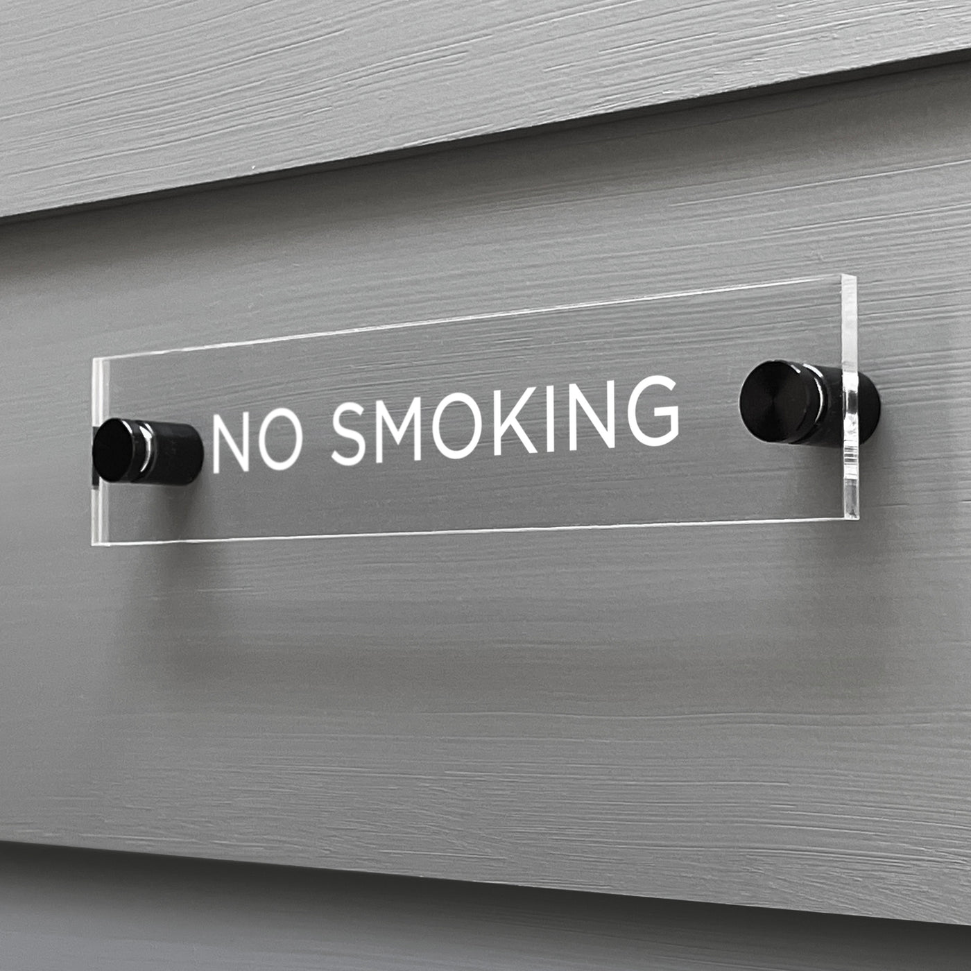 The No Smoking Acrylic Sign by Girl Friday Shop features sleek, clear acrylic with bold white letters and is mounted on two black standoffs for a modern, minimalist look.