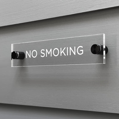 The No Smoking Acrylic Sign by Girl Friday Shop features sleek, clear acrylic with bold white letters and is mounted on two black standoffs for a modern, minimalist look.
