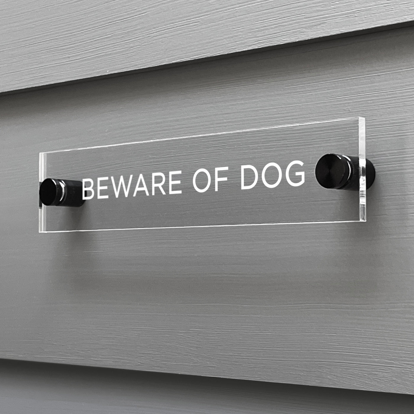 On a gray wall, a custom Girl Friday Shop Baware of Dogs Acrylic Sign displays the warning in bold white letters. Mounted with black brackets, this sign effectively conveys its message.