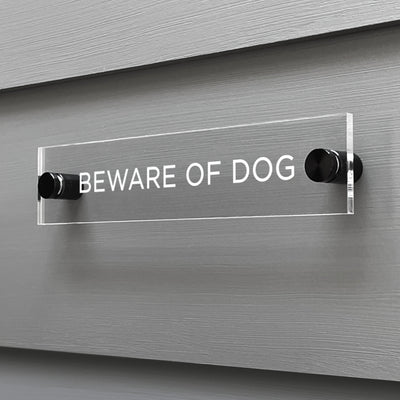 On a gray wall, a custom Girl Friday Shop Baware of Dogs Acrylic Sign displays the warning in bold white letters. Mounted with black brackets, this sign effectively conveys its message.