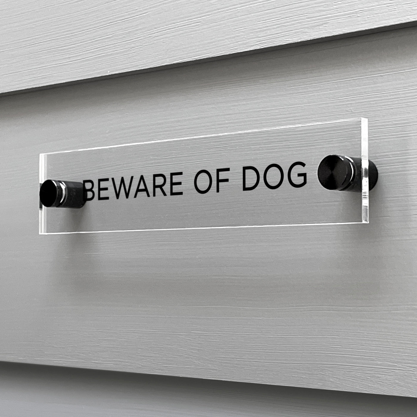 The Girl Friday Shops Baware of Dogs Acrylic Sign adorns a gray wall, displaying bold black capital letters, BEWARE OF DOG, mounted securely with two silver fasteners.