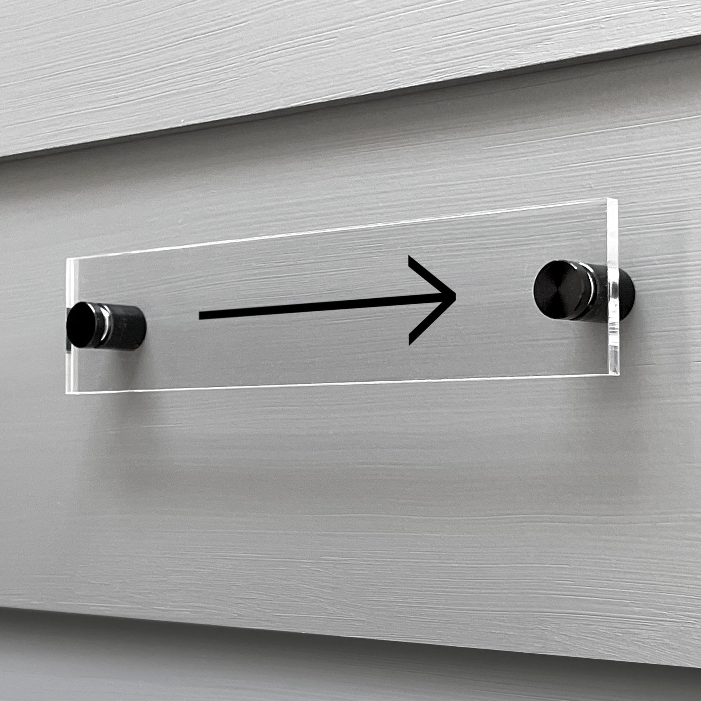 The Arrow Acrylic sign by Girl Friday Shop is mounted on a gray wall with two silver bolts, showcasing a sleek black arrow pointing to the right.