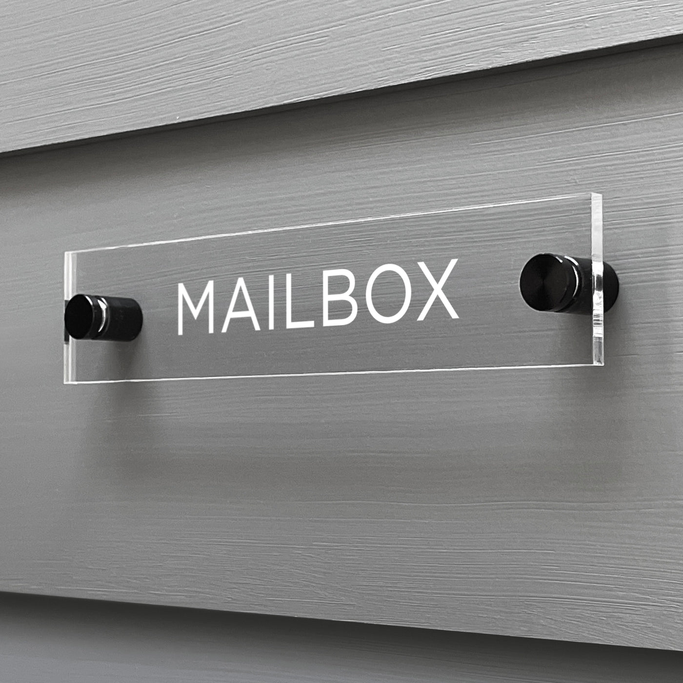 The Mailbox Acrylic Sign by Girl Friday Shop, featuring bold white letters, is custom-designed and elegantly mounted on a gray wooden wall with two cylindrical metal posts.
