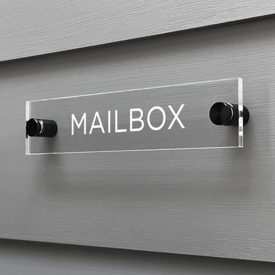 The Mailbox Acrylic Sign by Girl Friday Shop, featuring bold white letters, is custom-designed and elegantly mounted on a gray wooden wall with two cylindrical metal posts.