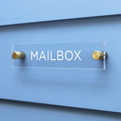 The Girl Friday Shop Mailbox Acrylic Sign features bold white capital letters on a clear surface, mounted on a light blue wall using two gold screws.